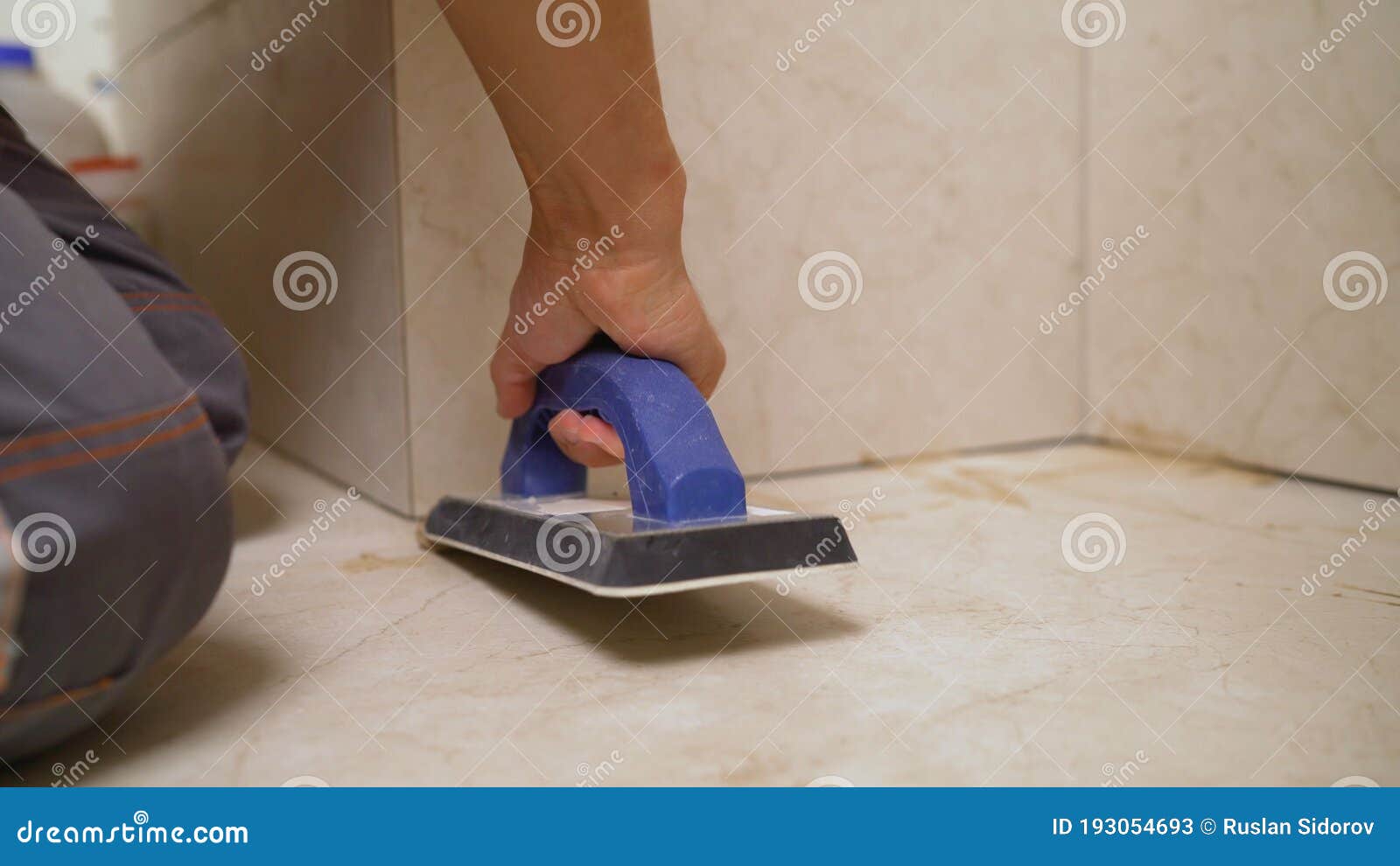 the hand of man holding a rubber float and filling joints with grout. the worker is rubbing the tiles with a float