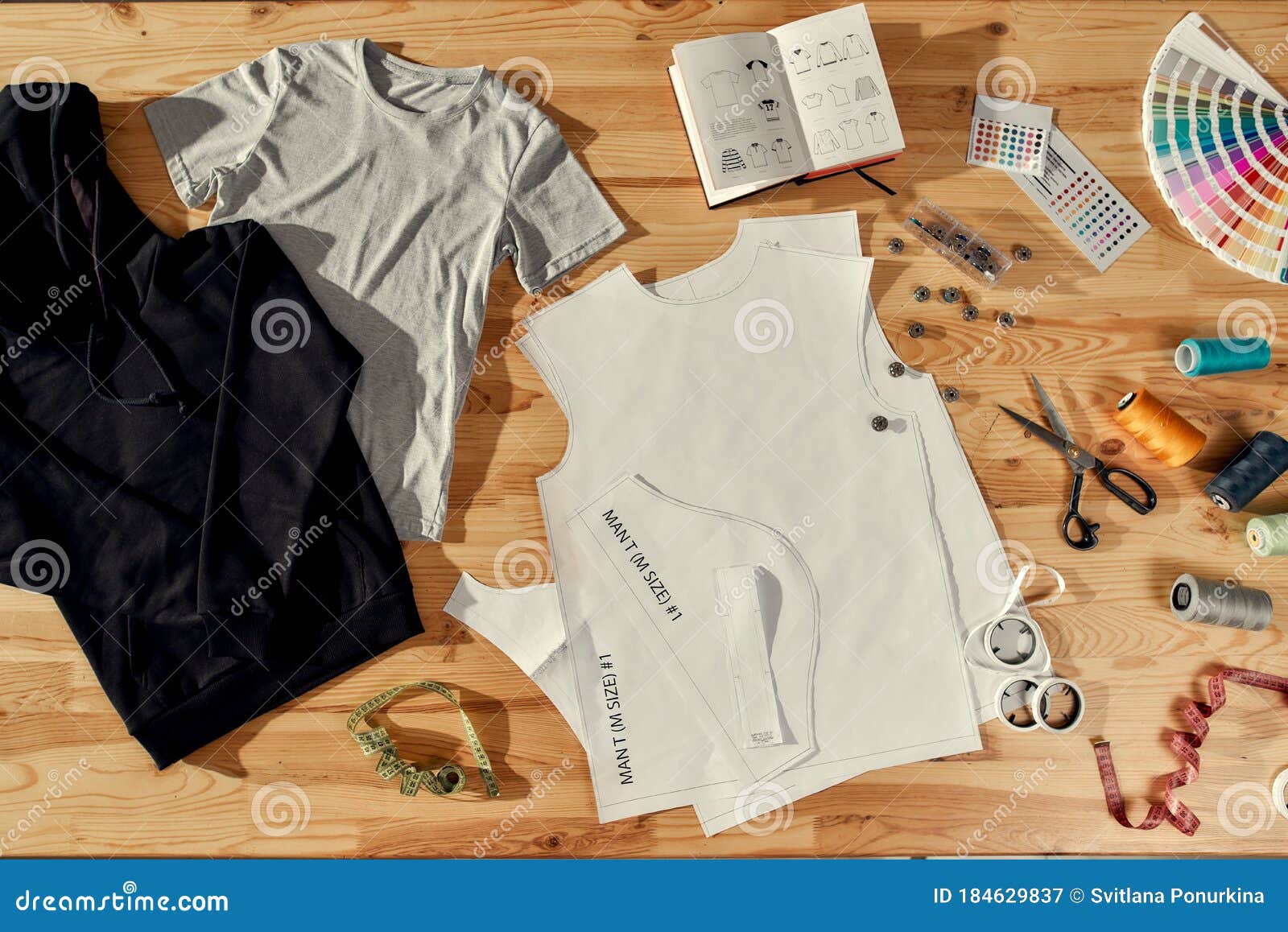 Workspace for Designer. Top view of neatly folded custom t shirts and  arranged baseball caps. Samples of fabric, stickers with text, tape measure  and notebooks lying on a wooden background. Stock Photo