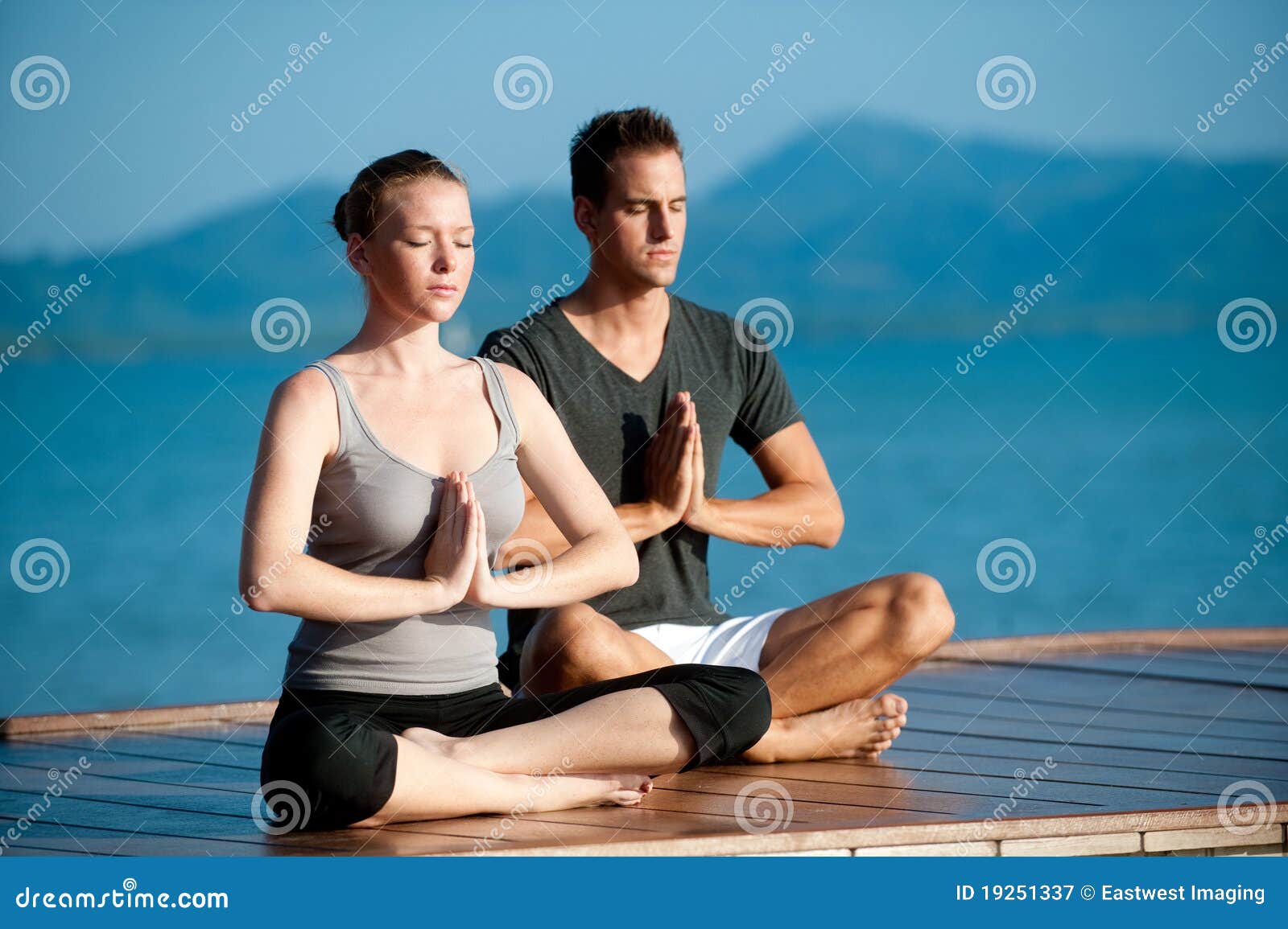 Yogapar av Hav. En attraktiv ung kvinna och man som gör yoga på en brygga med det blåa hav och en annan ö bak dem