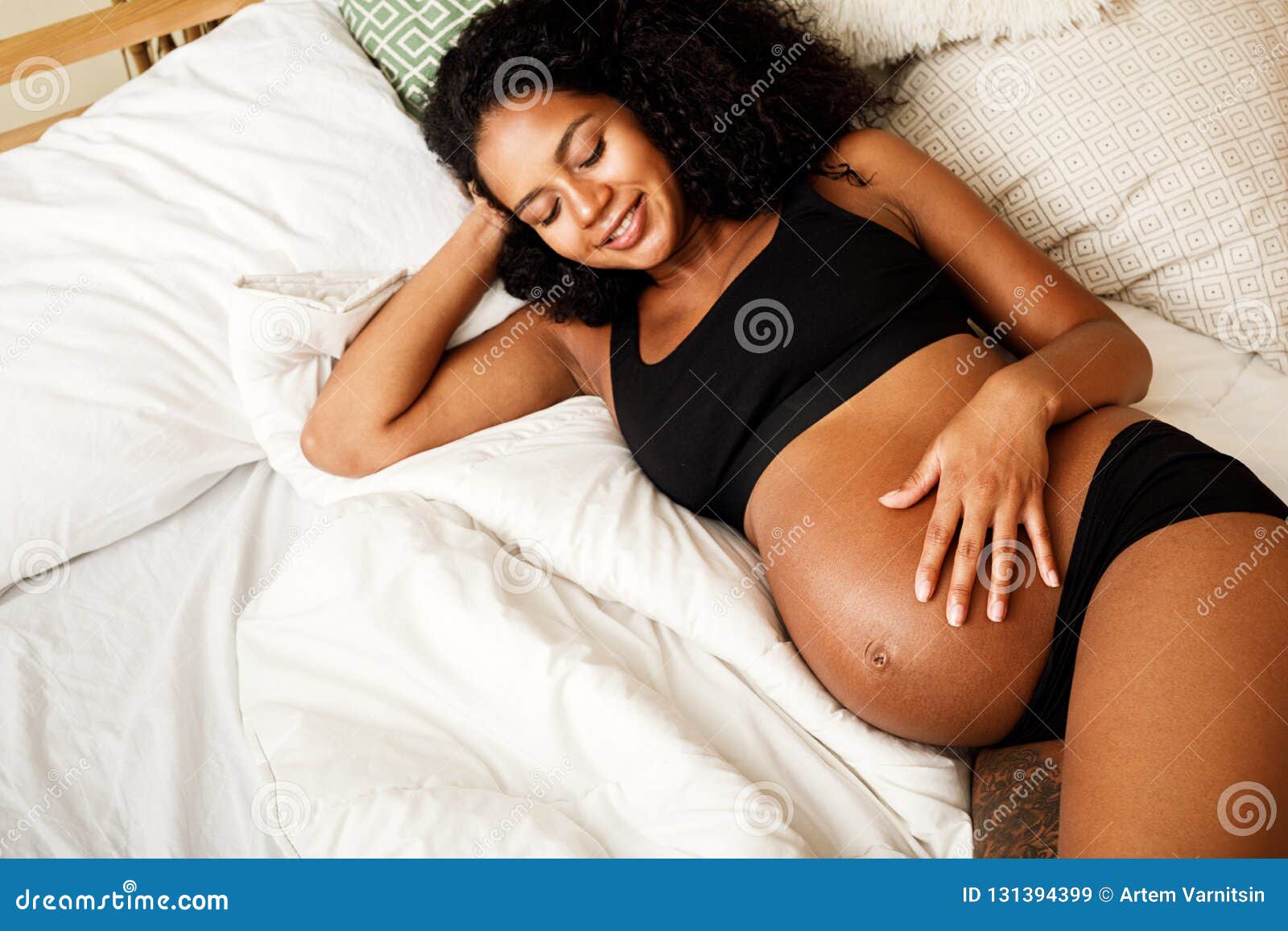 Foto de Portrait of beautiful young African American woman in underwear  lying relaxing on white bed at home. Smiling pretty millennial black female  in underclothing garment. Body positive concept. do Stock
