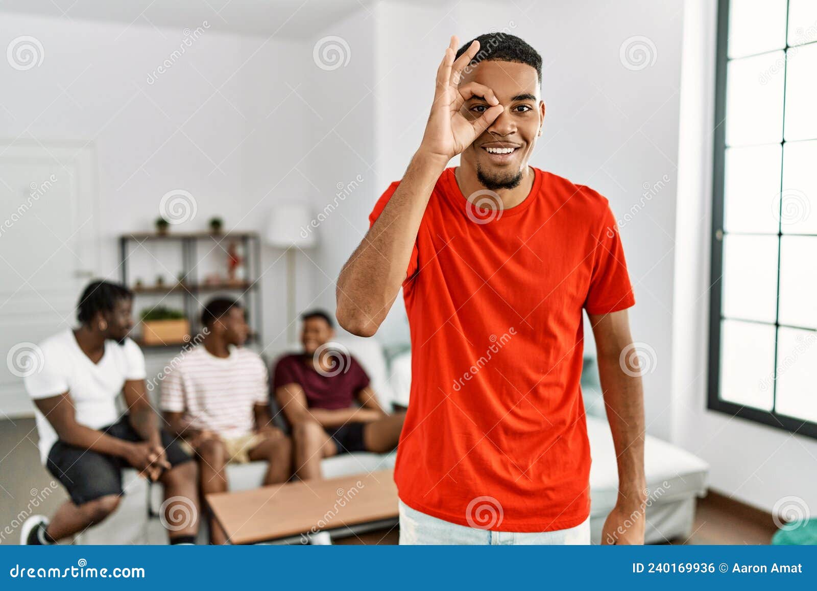 young african man at home with friends sitting on the sofa at home smiling happy doing ok sign with hand on eye looking through