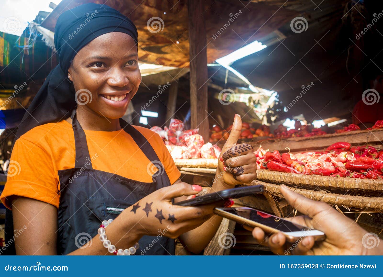 pretty young black woman selling tomatoes in a local african market receiving payment via mobile phone transfer giving a thumbs up