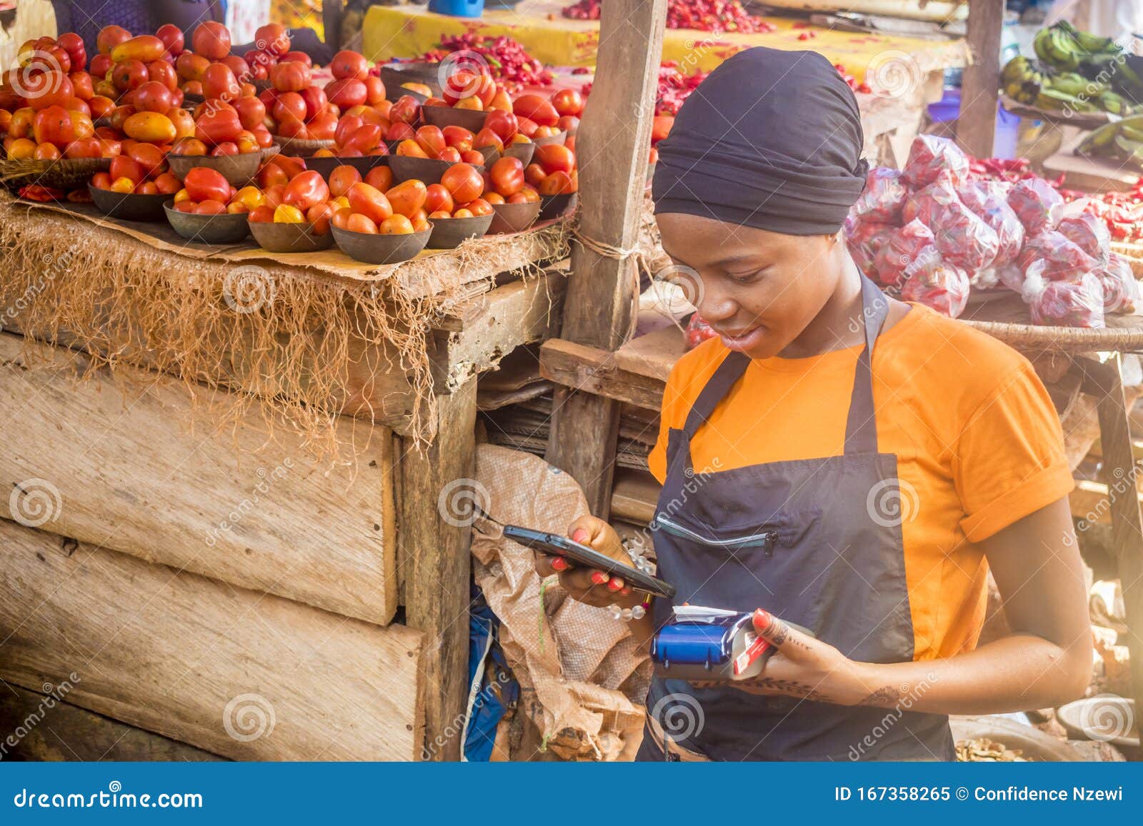 young african woman selling tomatoes in a local african market using her mobile phone and holding a mobile pos device.