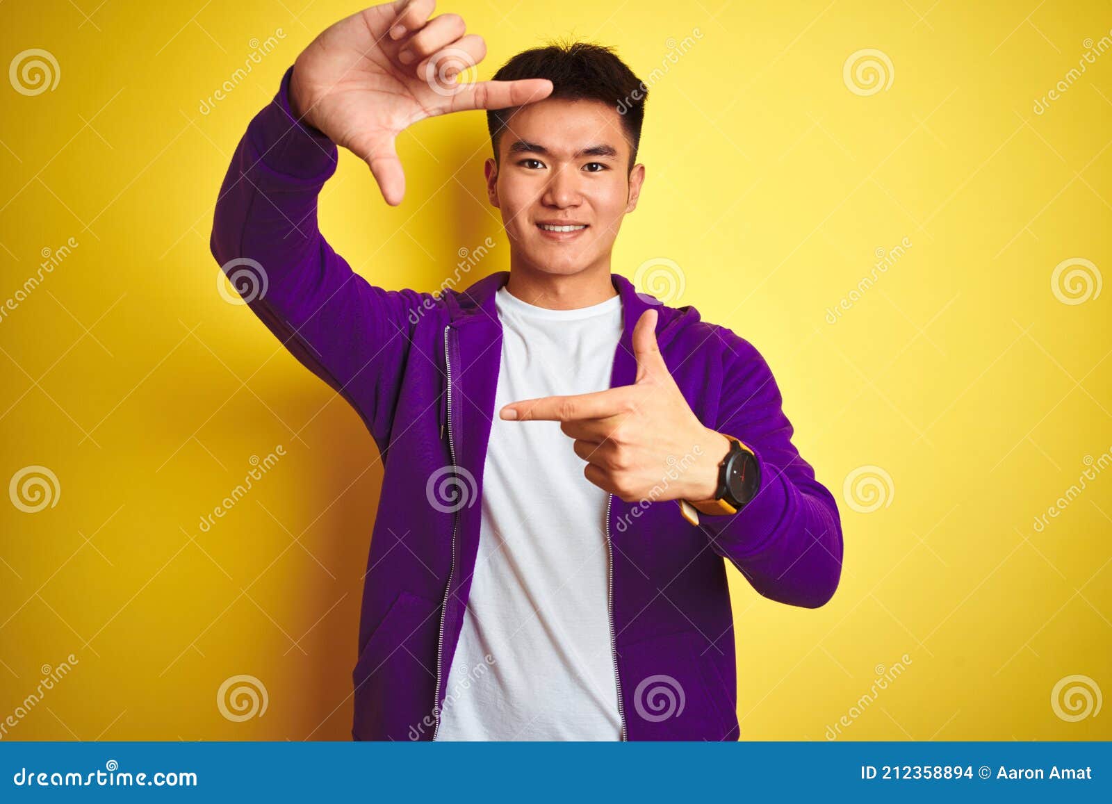 Young asian chinese man wearing purple sweatshirt standing over isolated yellow background smiling making frame with hands and fingers with happy face. Creativity and photography concept