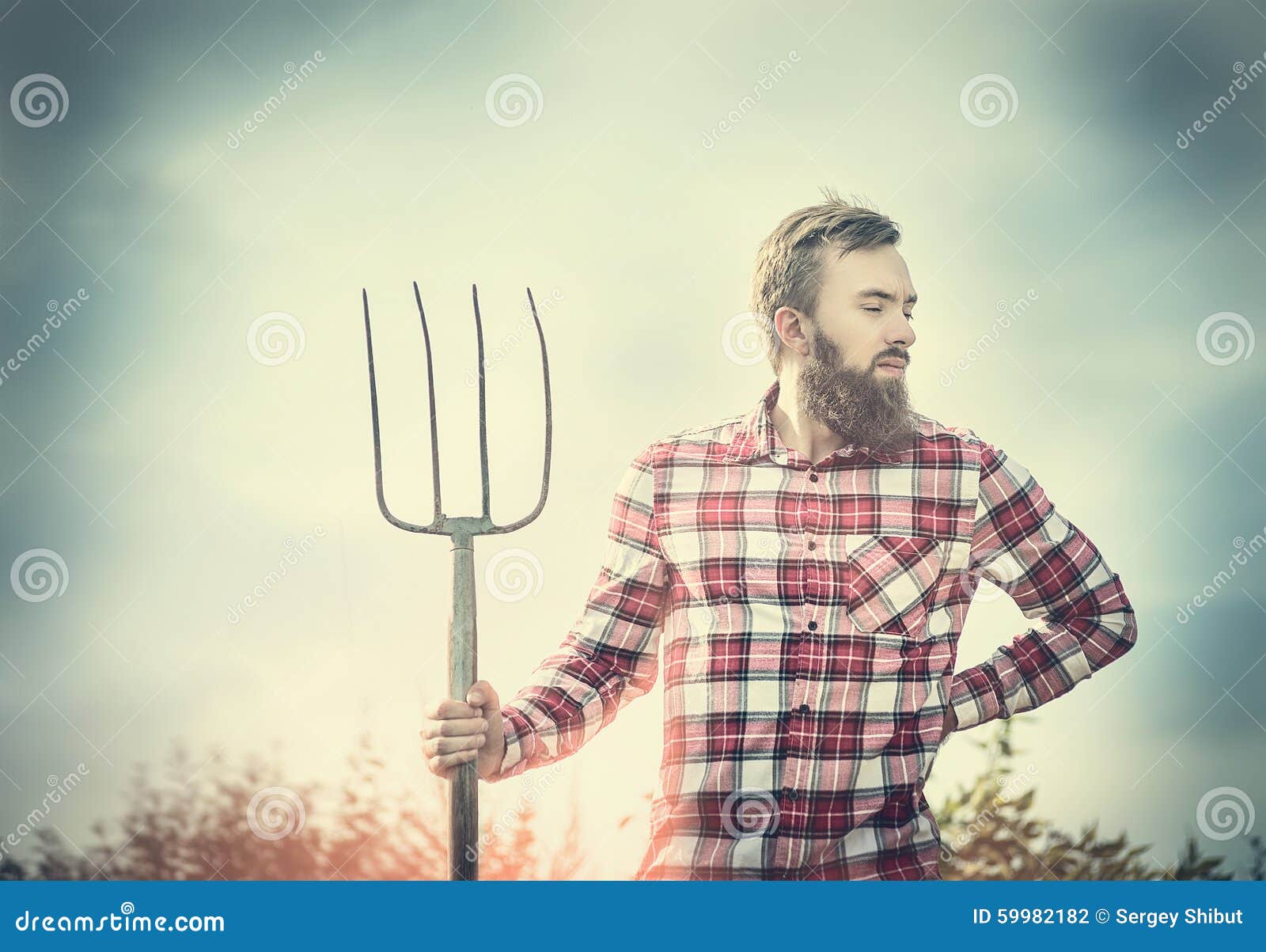 young bearded farmer in red checkered shirt with old pitchfork sky nature backgrund, toned