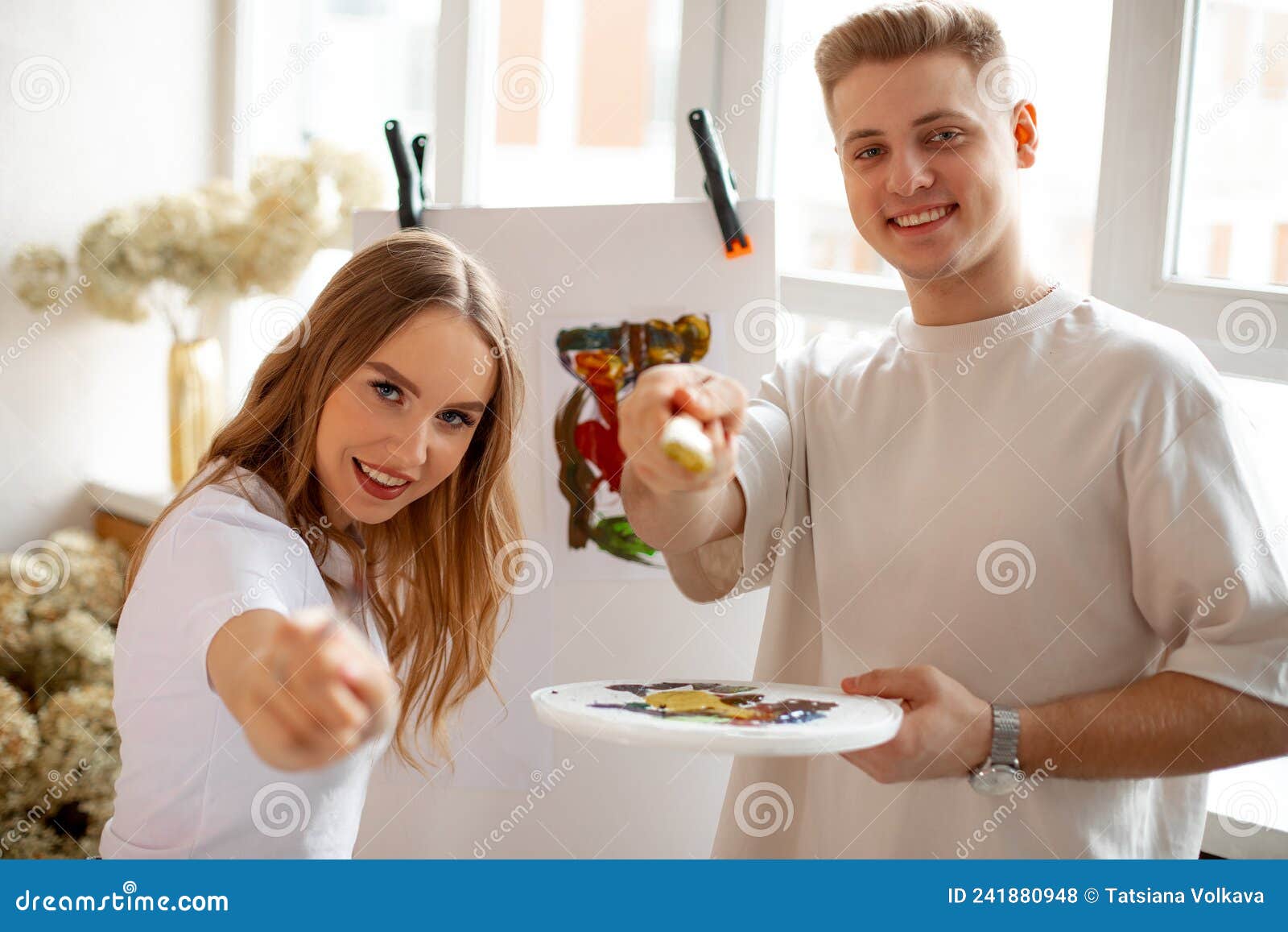 young beauteous couple stand pointing with brushes at camera while man holding watercolor palette in front of canvas.