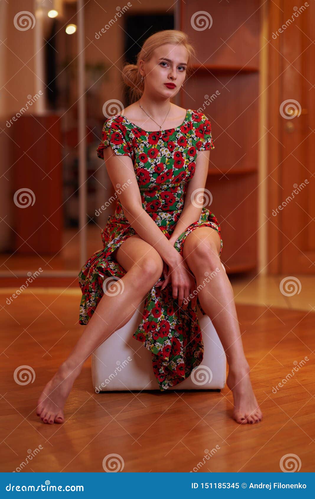 a young beautiful blonde with hair gathered in a chignon in a floral print dress sits on a ottoman in a home setting, pulling off
