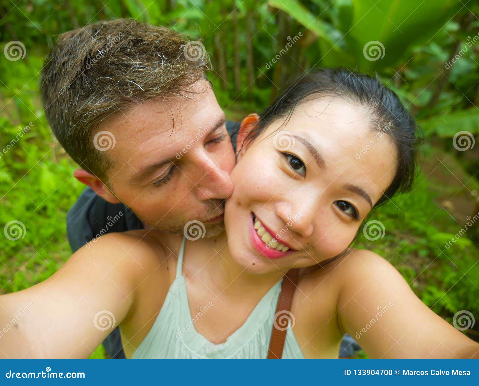 Young beautiful and happy mixed ethnicity couple beautiful Asian Chinese woman and white man in love taking selfie picture. Young beautiful and happy mixed ethnicity couple beautiful Asian Chinese women and white men in love taking selfie picture outdoors enjoying romantic holidays trip in tropical honeymoon vacation