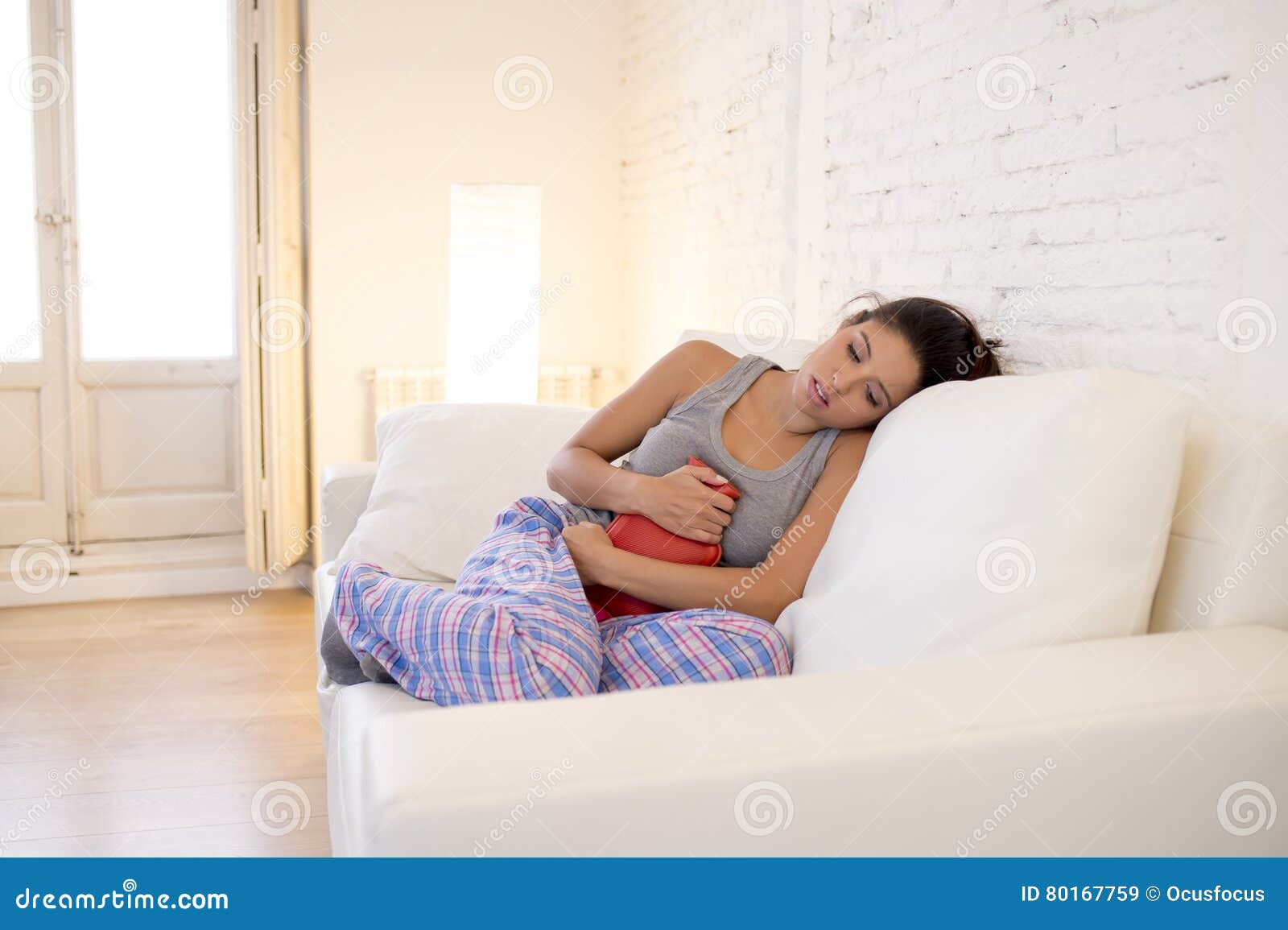 young beautiful hispanic woman holding hot water bottle against belly suffering menstrual period pain