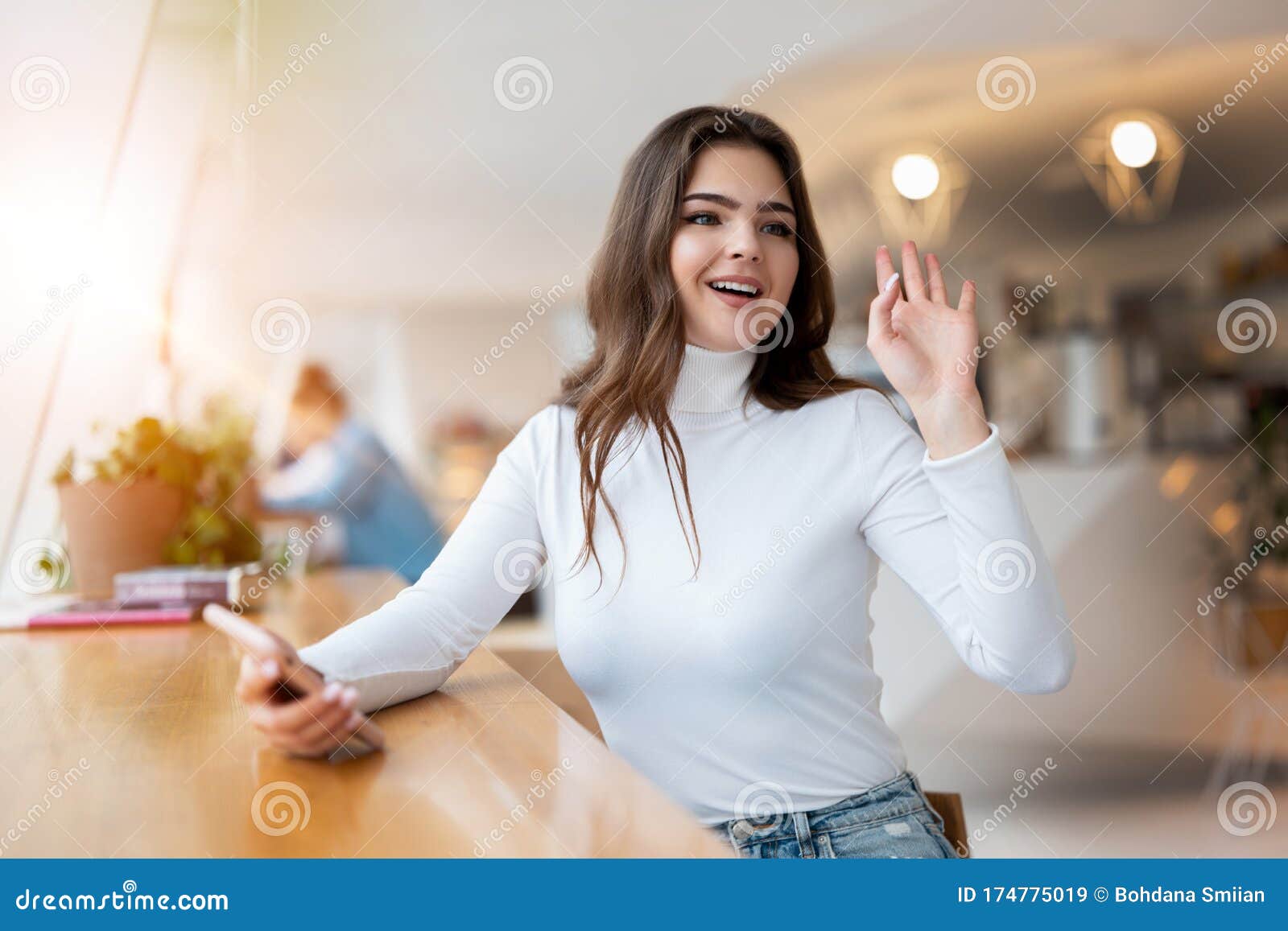 young beautiful woman with smarphone in her hand waving her friend l while sitting in the cafe during lunch break, positive vibes