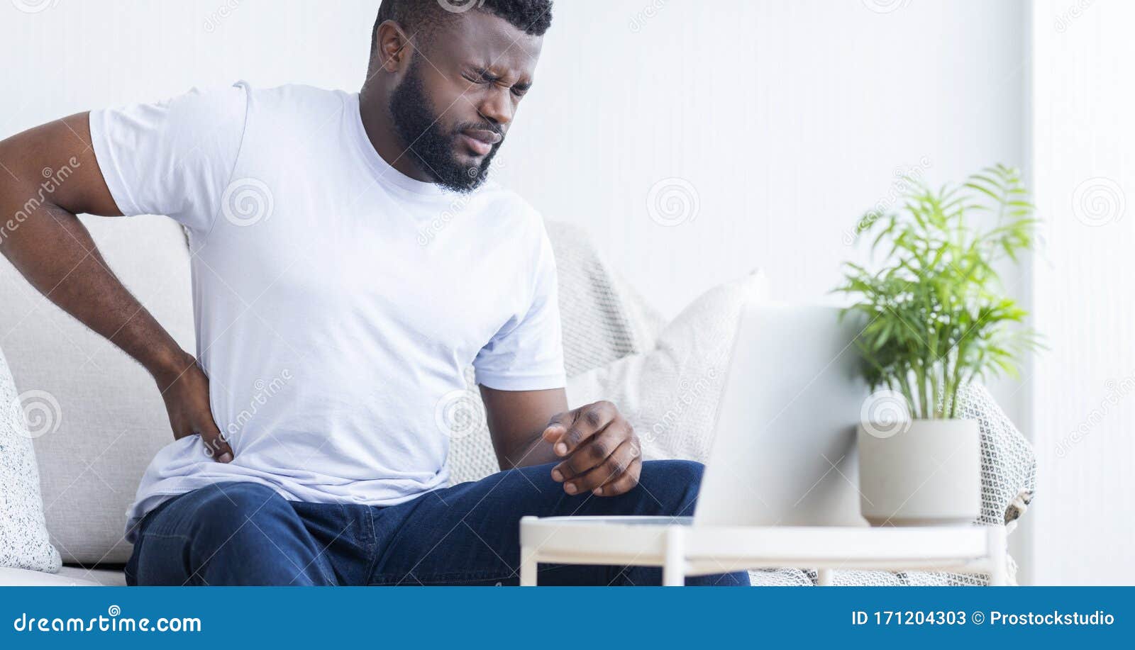 young black man touching his sore back, working on laptop
