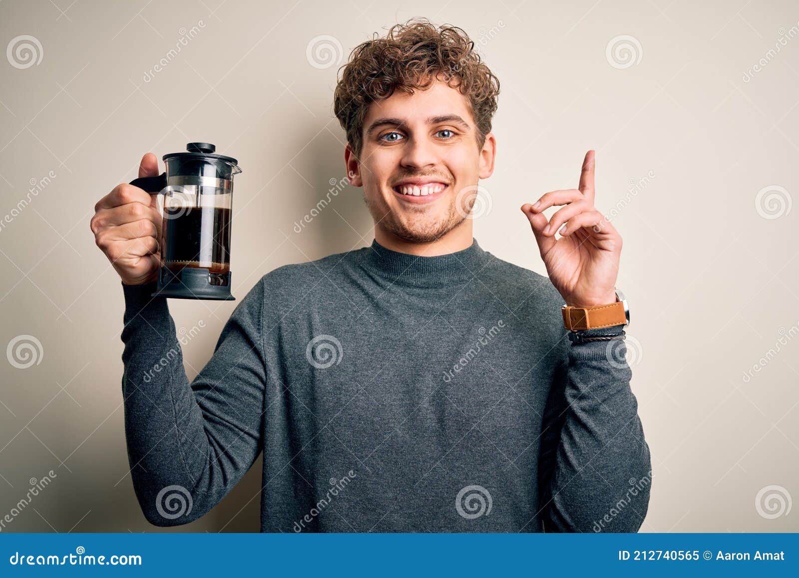 young blond man with curly hair making coffee using coffemaker over white background surprised with an idea or question pointing