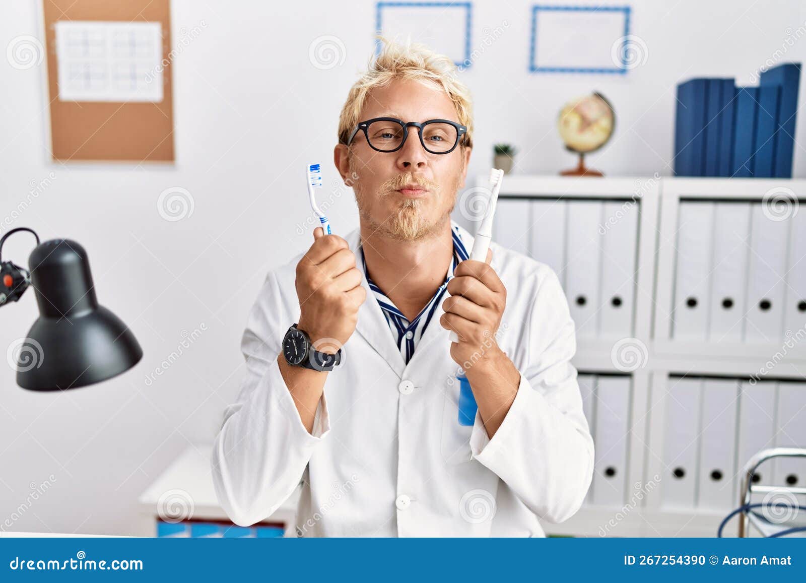 young blond man working at dentist clinic holding electric toothbrush and teethbrush looking at the camera blowing a kiss being