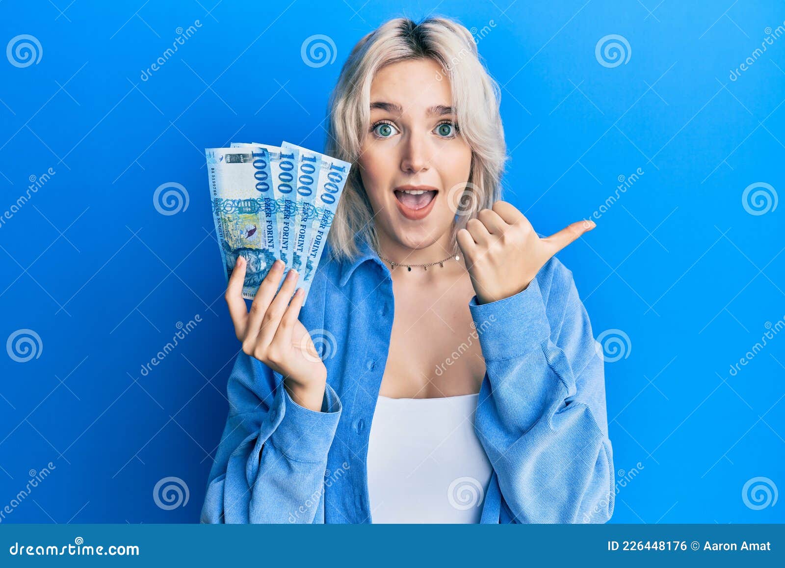 Young Blonde Girl Holding Hungarian Forint Banknotes Pointing Thumb Up To The Side Smiling Happy