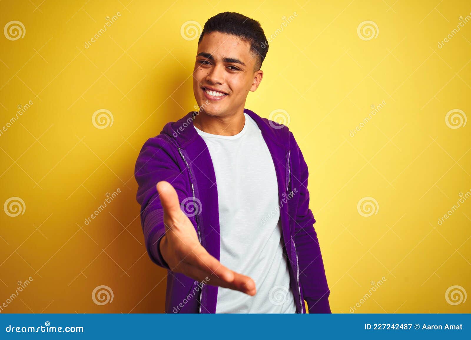 young brazilian man wearing purple sweatshirt standing over  yellow background smiling friendly offering handshake as