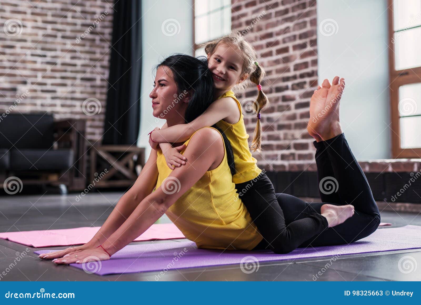 young caucasian mother doing yoga cobra pose on floor while her smiling daughter sitting on moms back hugging her in