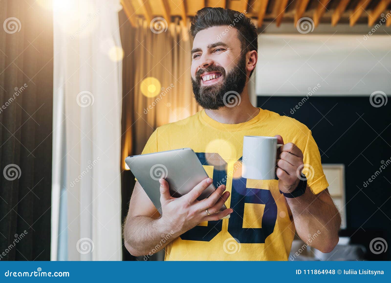young bearded man stands in room and holding tablet computer while drinking coffee. guy freelancer working at home.