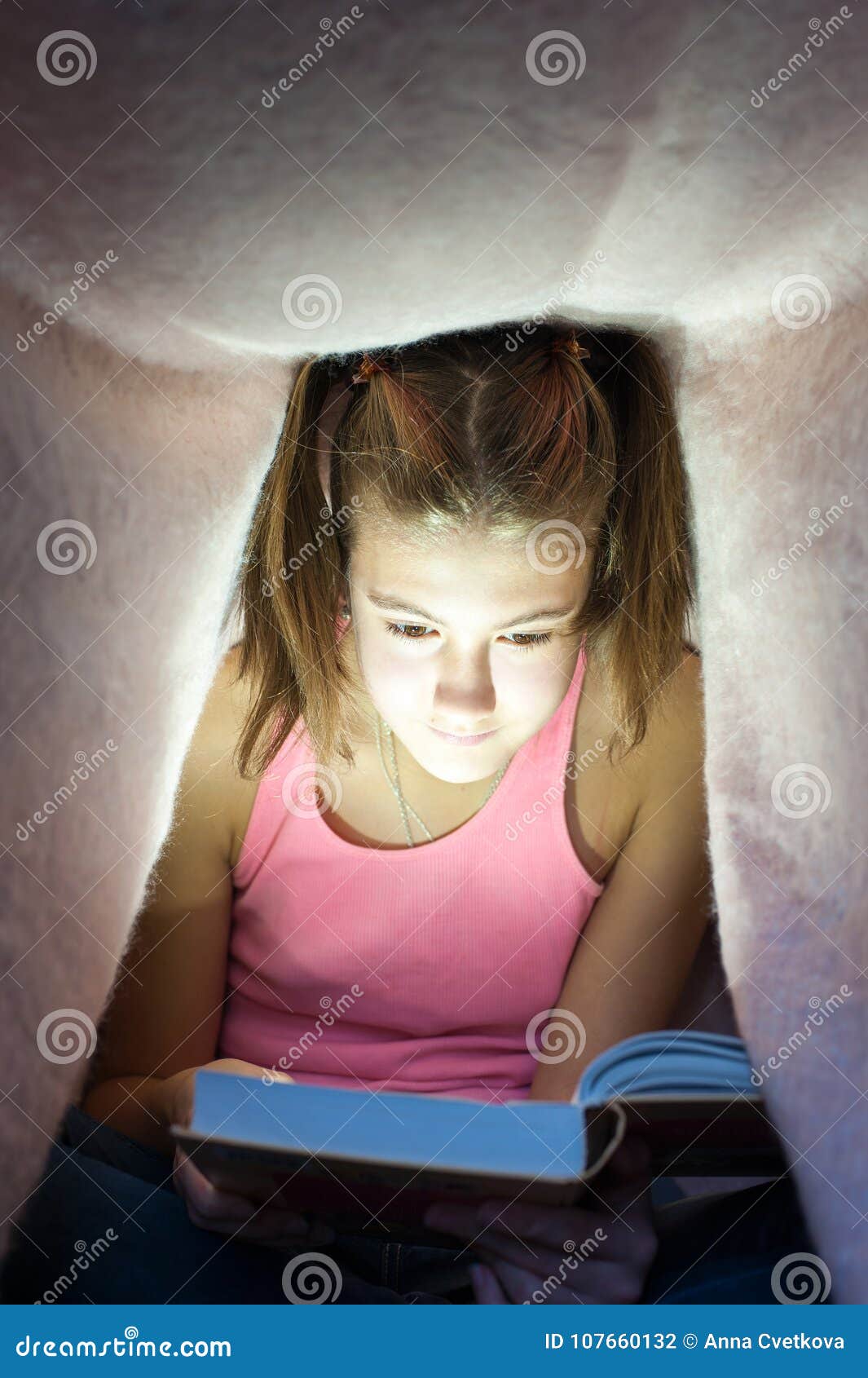 young cheerful girl hiding under blanket and enrapt reading book
