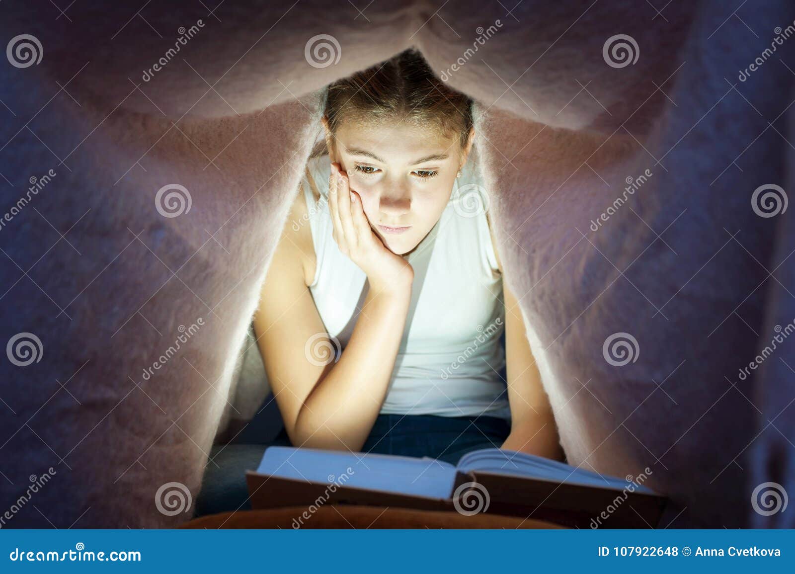 young cheerful girl hiding under blanket and enrapt reading book