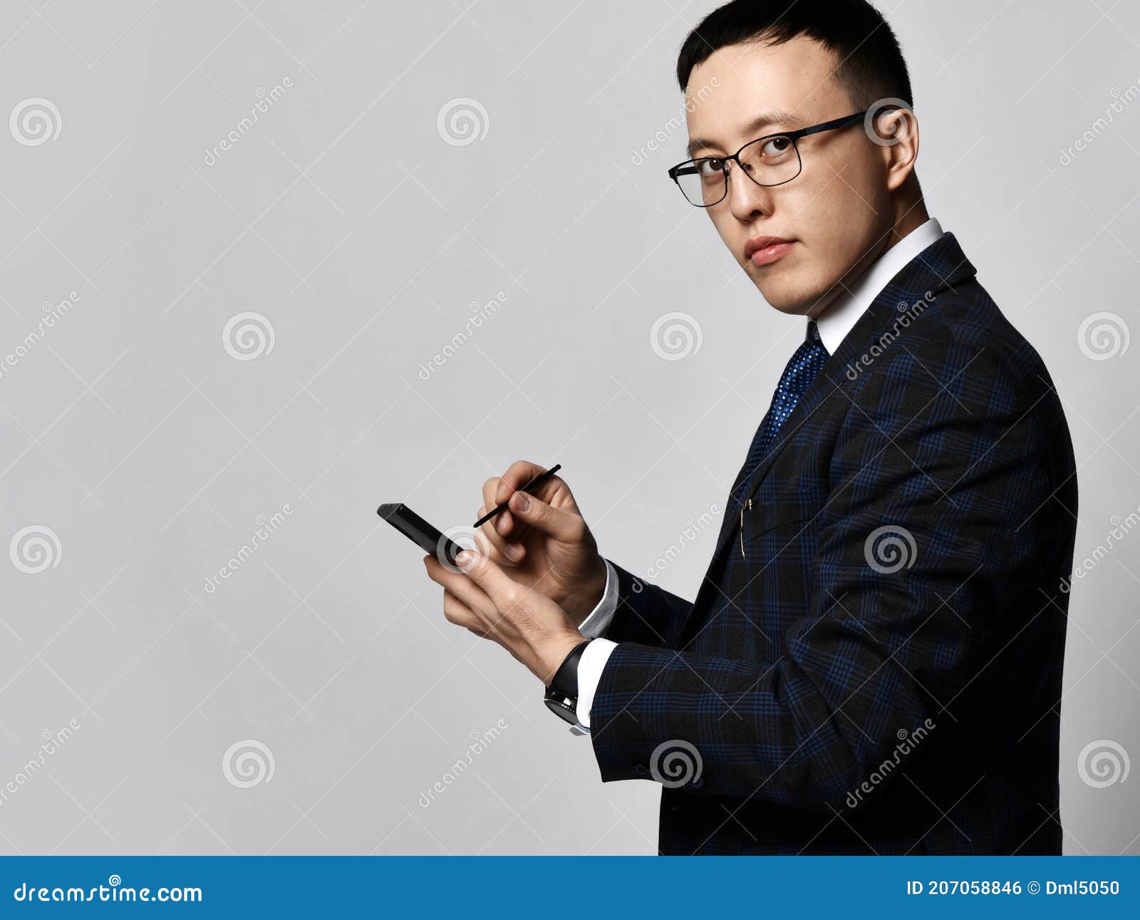 young confident asian businessman in checkered official suit, tie and glasses standing and making notes in smartphone
