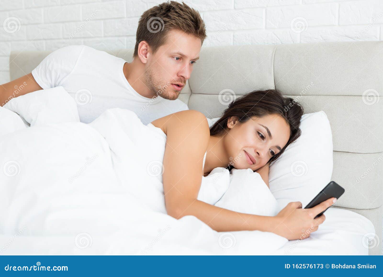 Young Couple Handsome Husband and His Beautiful Wife Just Woke Up in Bed Woman Checking Her Smartphones Early in the Stock Image photo