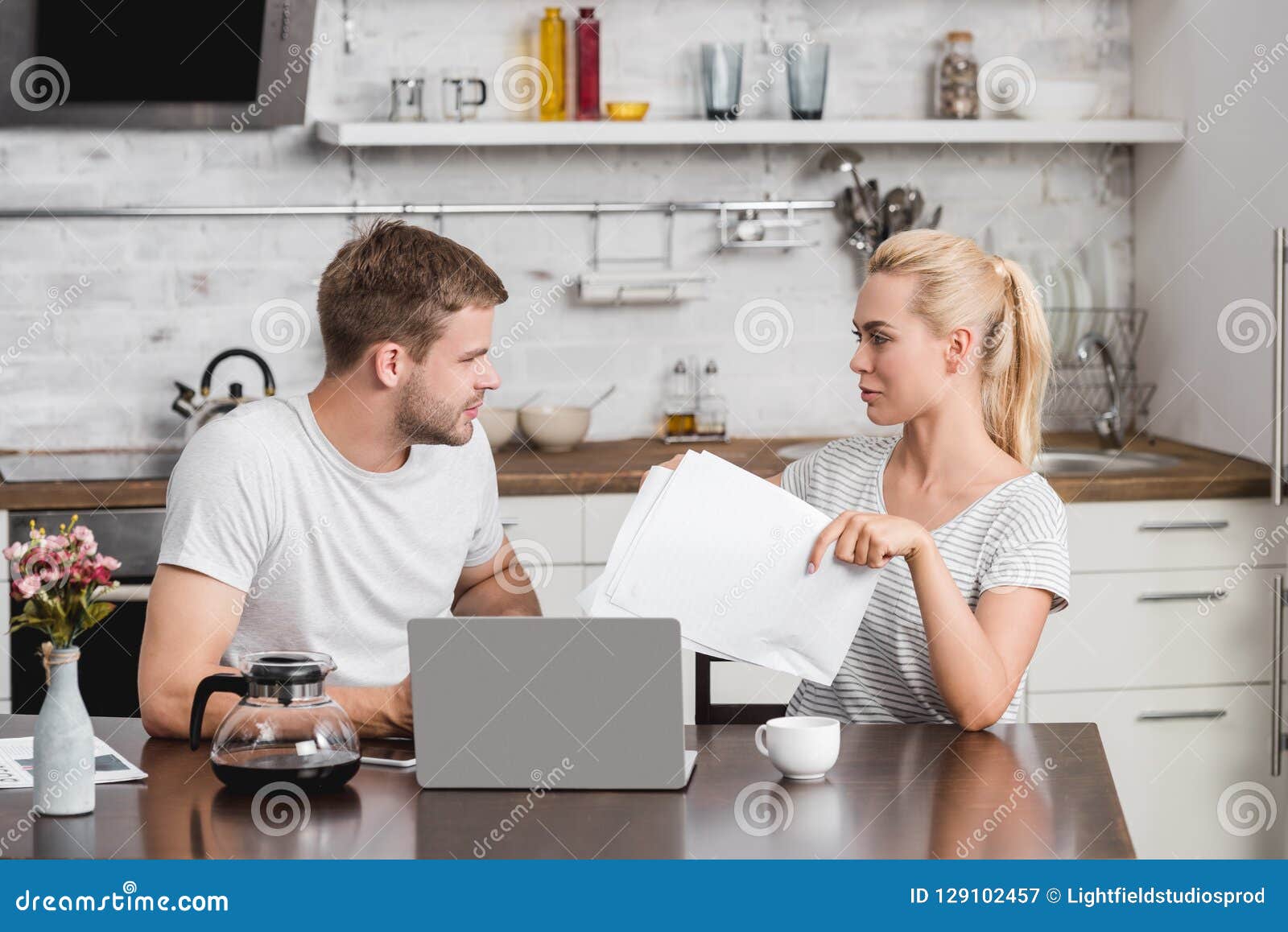 young couple holding papers and using laptop while sitting at kitchen table and looking