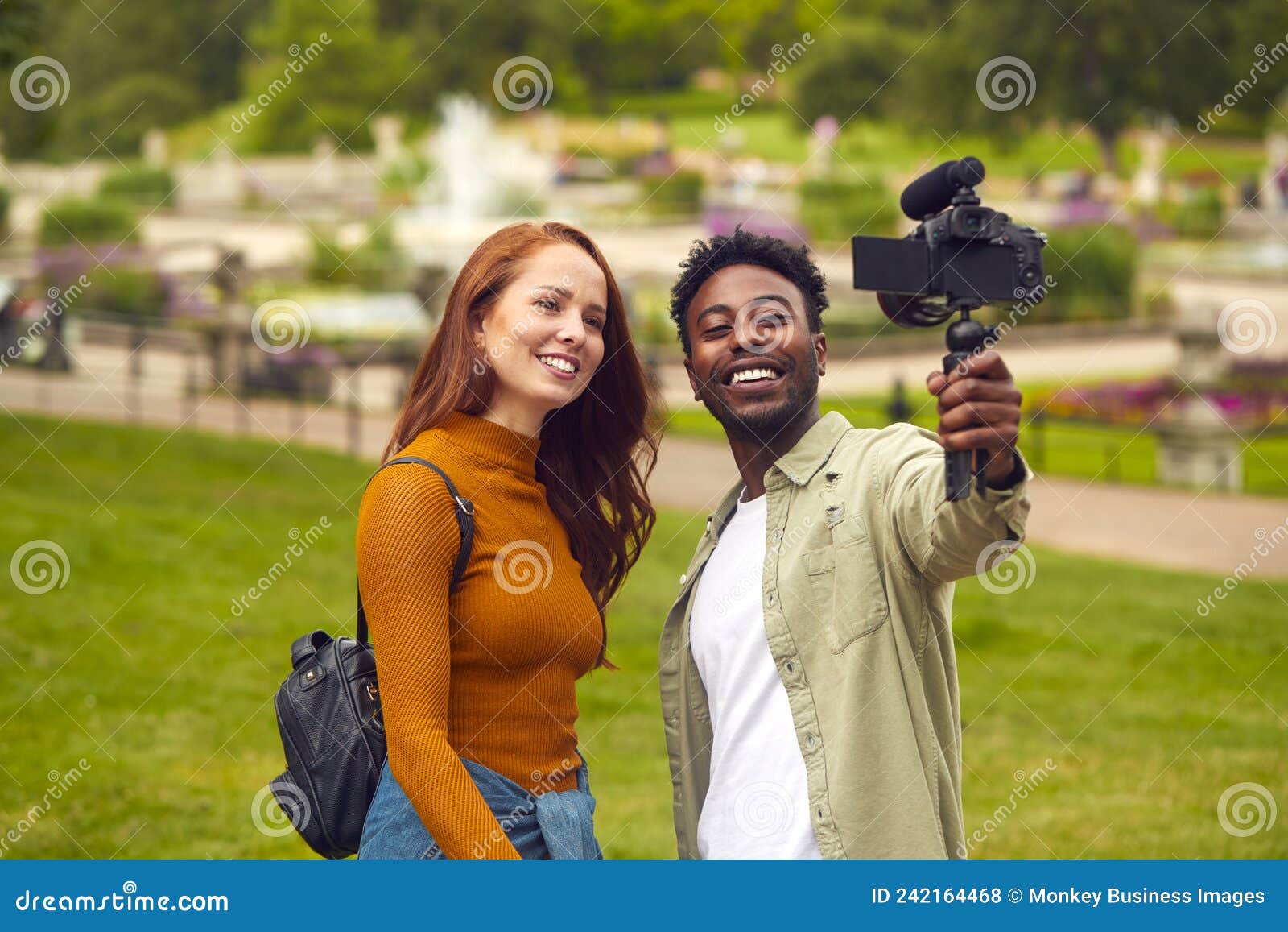 young couple travelling through city park together vlogging to video camera on handheld tripod