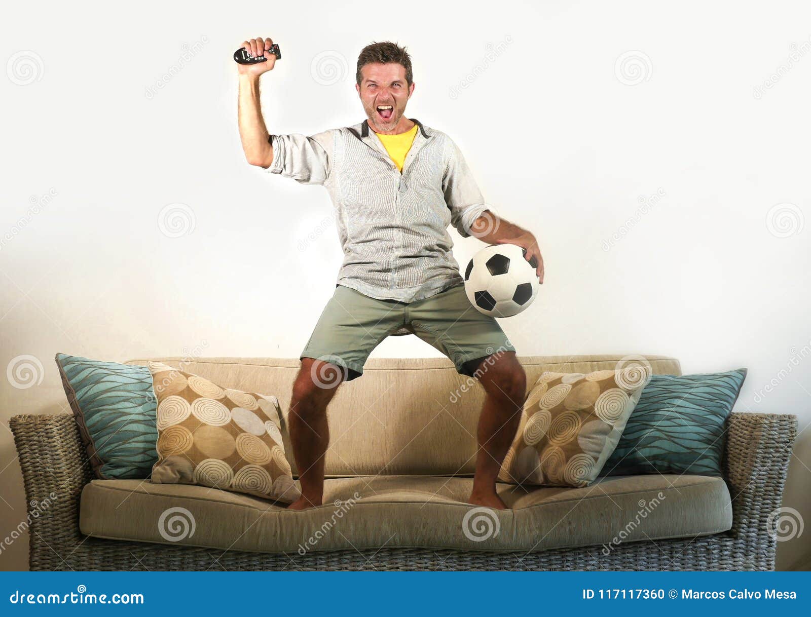 Young Enthusiastic Football Fan Celebrating Goal Crazy Happy Jumping on Sofa Couch at Home Watching Soccer Game on Television Hold Stock Photo