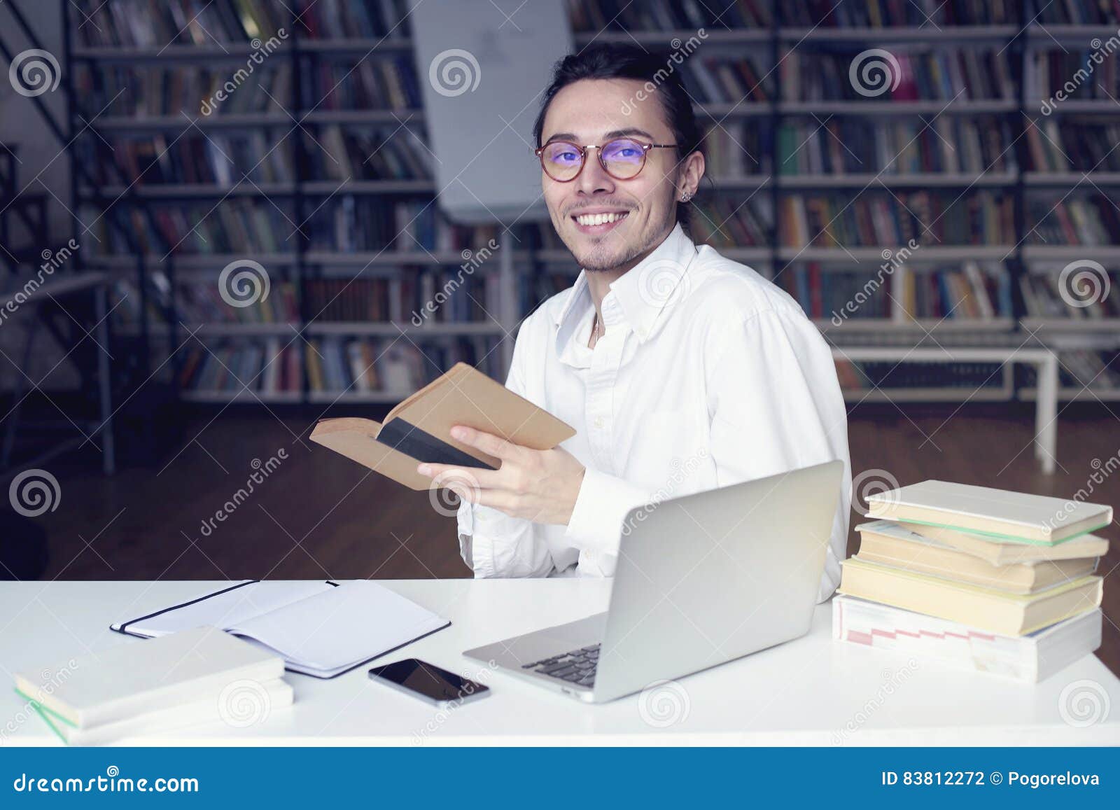 young entrepreneur or university student smiling, working on laptop reading a book in a library