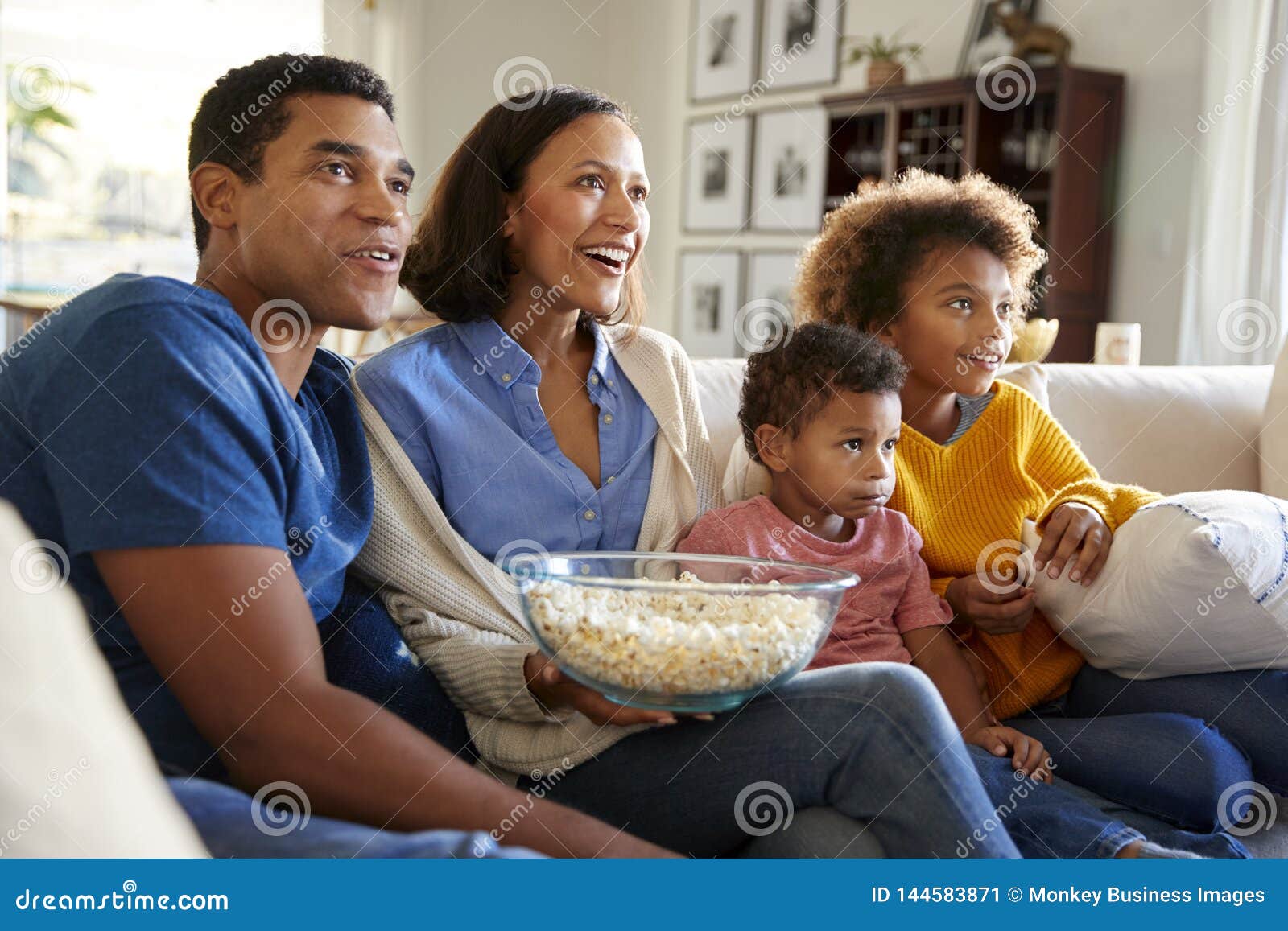 young family sitting together on the sofa in their living room watching tv and eating popcorn, side view