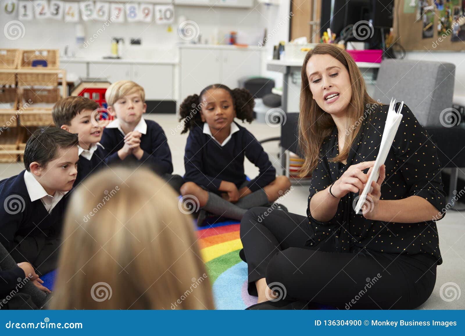young female primary school teacher reading a book to children sitting on the floor in a classroom, selective focus