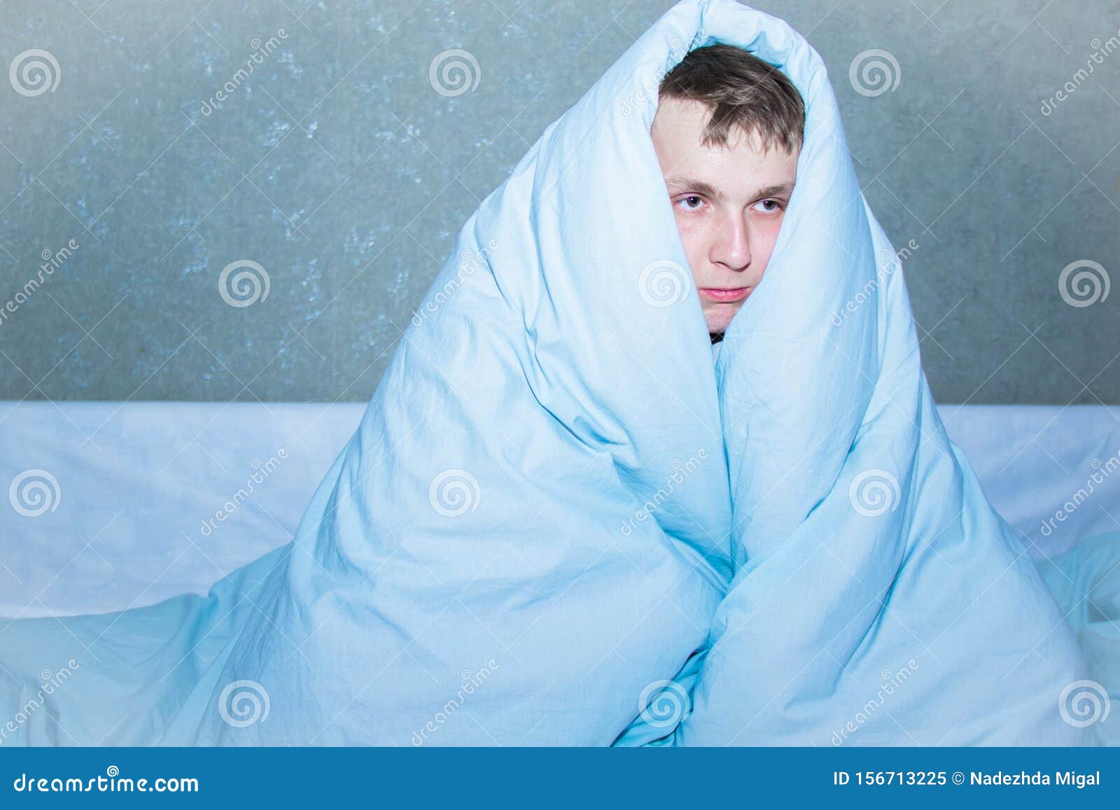 Young Guy Sitting on a Bed Wrapped in a Blue Blanket. Stock Image