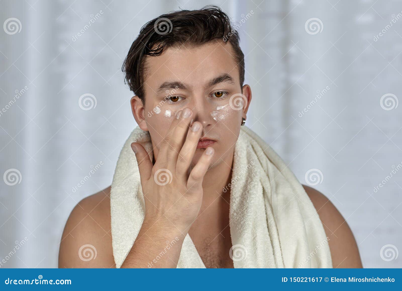 young handsome caucasian man applying cream under the eyes, towel on shoulders. caring face, metrosexual daily routine in the bath