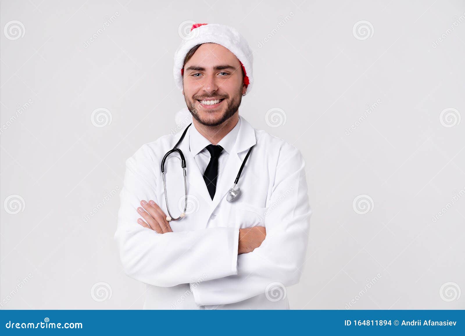 young handsome doctor in white uniforme and santa claus hat standing in studio on white background loking at camera abd teeth