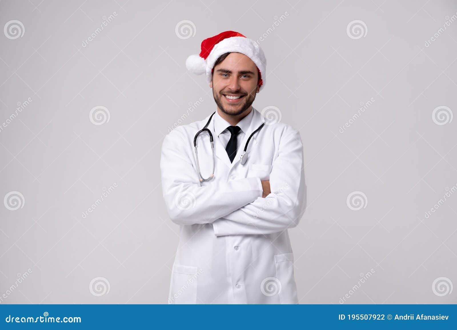 young handsome doctor in white uniforme and santa claus hat standing in studio on white background loking at camera abd teeth