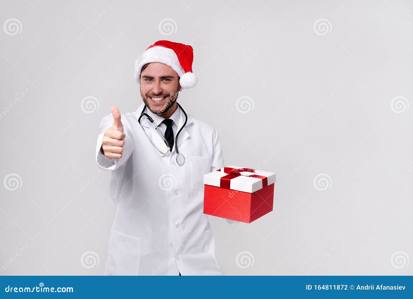 young handsome doctor in white uniforme and santa claus hat standing in studio on white background smile and finger in camera