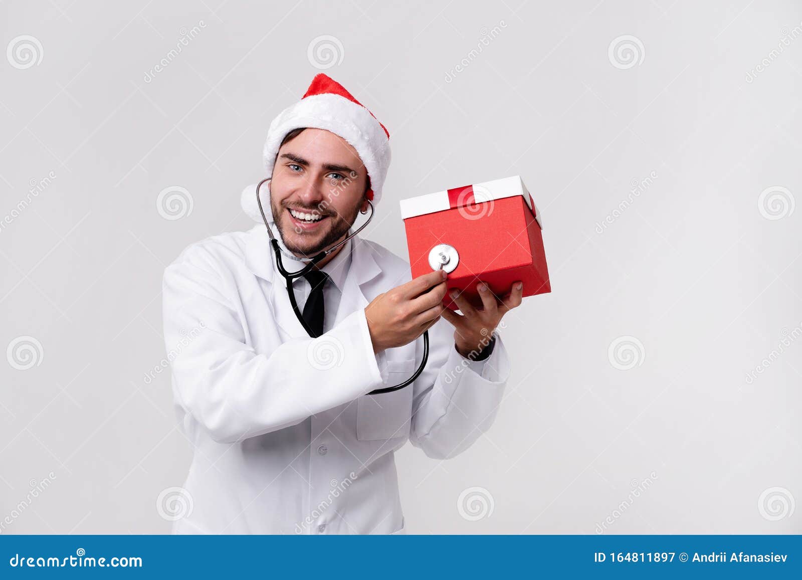 young handsome doctor in white uniforme and santa claus hat standing in studio on white background smile and finger in camera