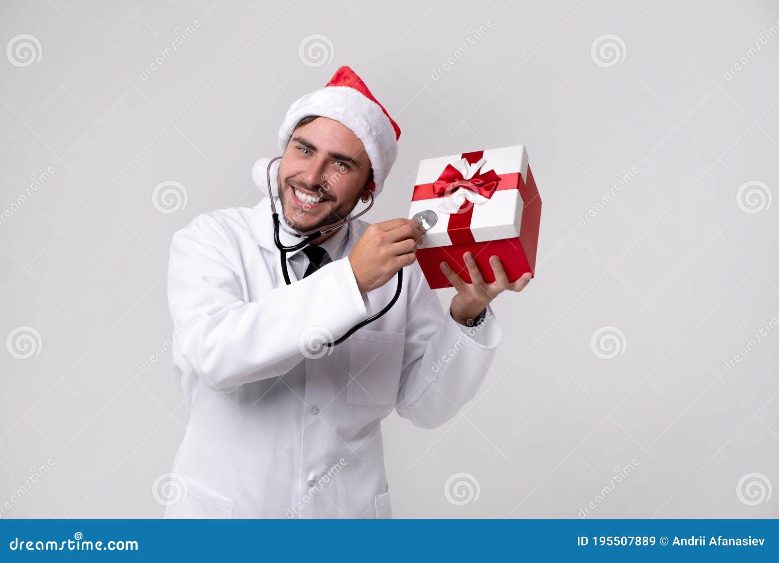 young handsome doctor in white uniforme and santa claus hat standing in studio on white background smile and finger in camera