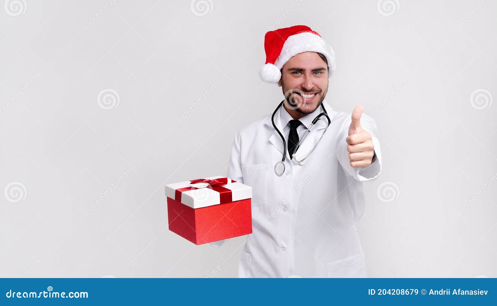 young handsome doctor in white uniforme and santa claus hat standing in studio on white background smile and finger in camera