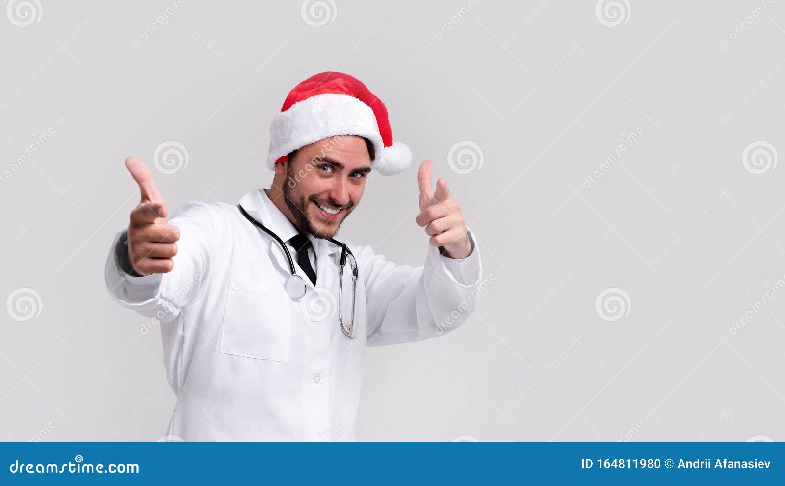 young handsome doctor in white uniforme and santa claus hat standing in studio on white background smile and finger in camera