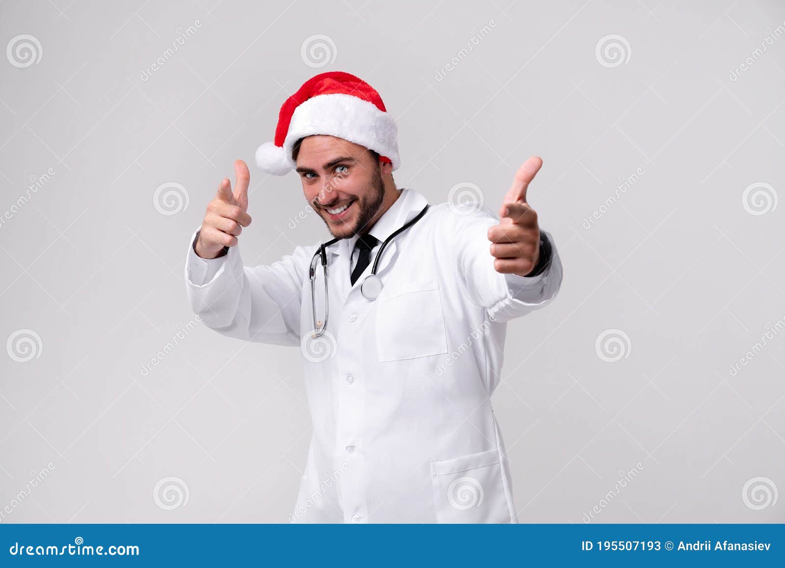 young handsome doctor in white uniforme and santa claus hat standing in studio on white background smile and finger in camera