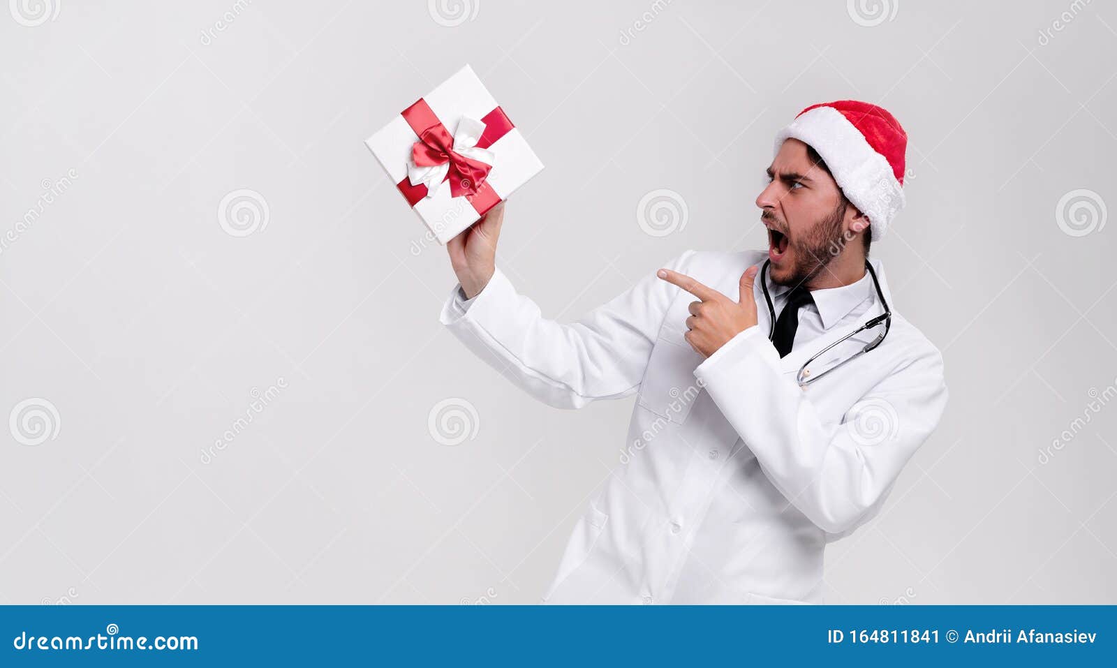 young handsome doctor in white uniforme and santa claus hat standing in studio on white background smile and shows a finger at the