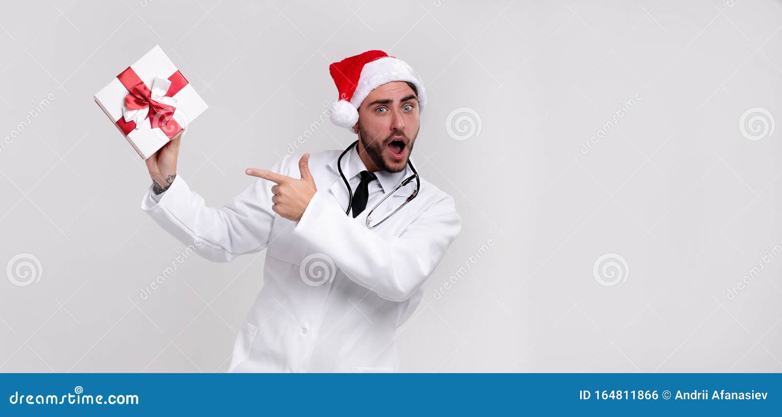 young handsome doctor in white uniforme and santa claus hat standing in studio on white background smile and shows a finger at the