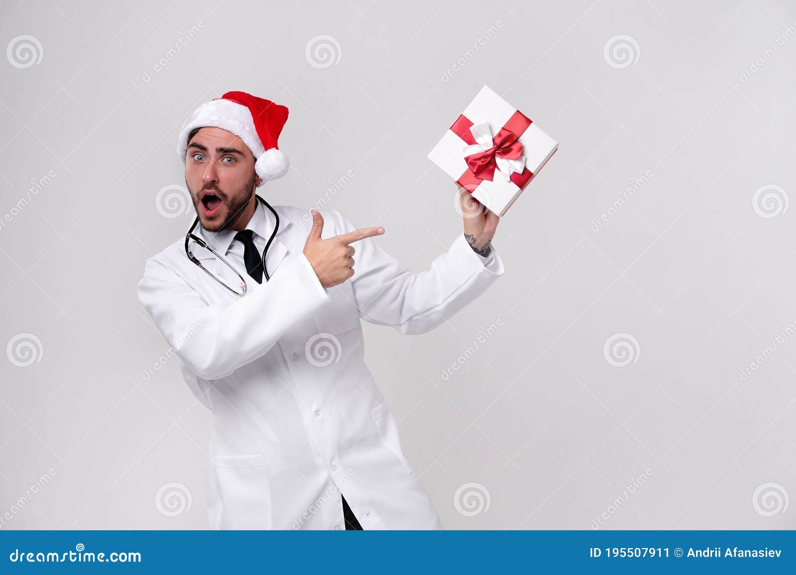 young handsome doctor in white uniforme and santa claus hat standing in studio on white background smile and shows a finger at the