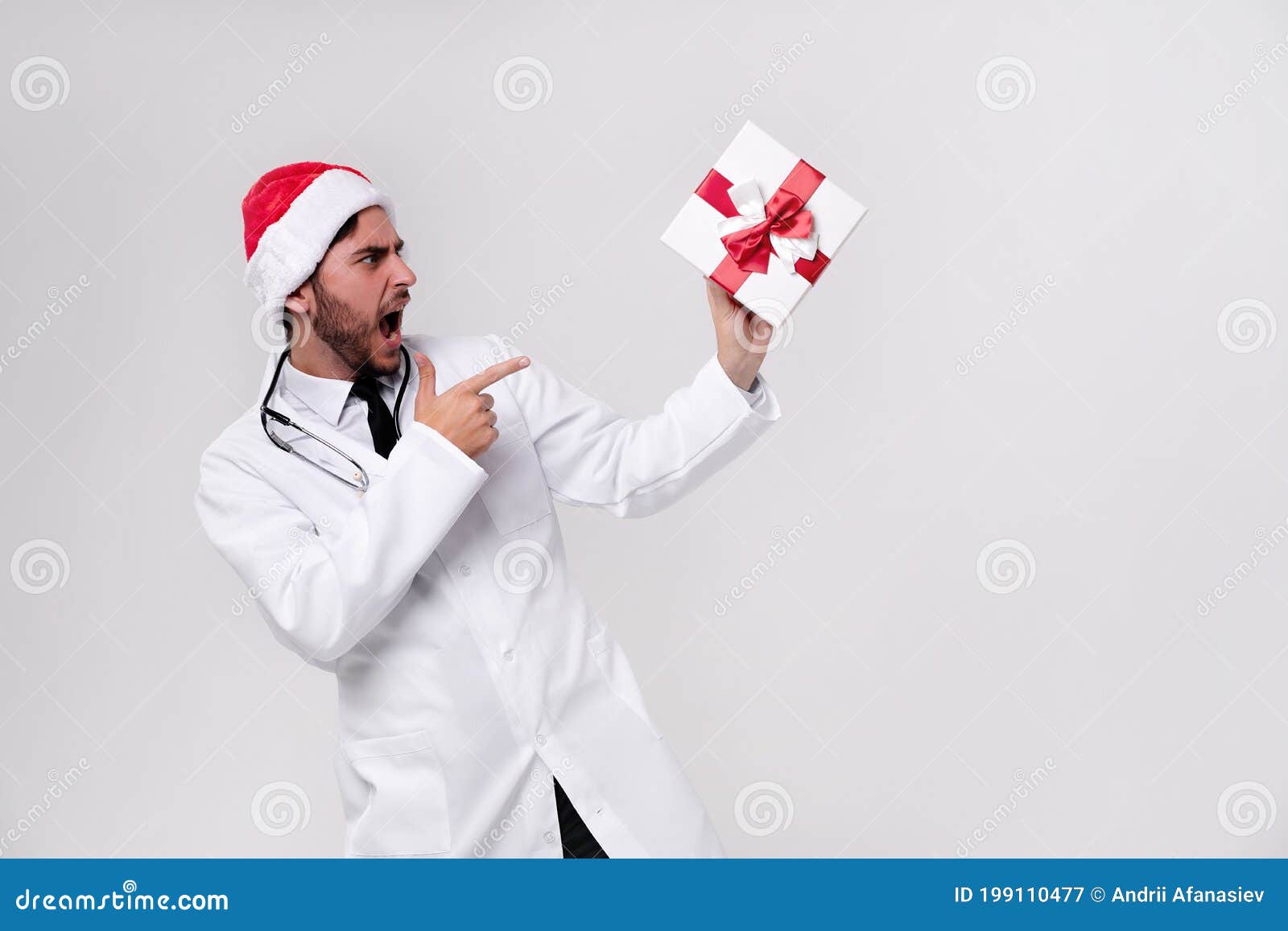 young handsome doctor in white uniforme and santa claus hat standing in studio on white background smile and shows a finger at the