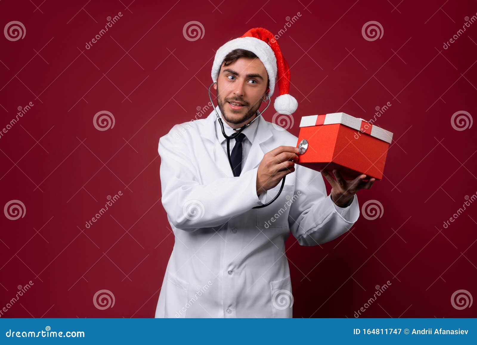 young handsome doctor in white uniforme and santa claus hat standing in studio on red background smile and finger in camera