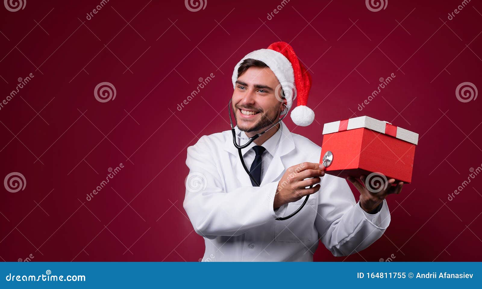 young handsome doctor in white uniforme and santa claus hat standing in studio on red background smile and finger in camera