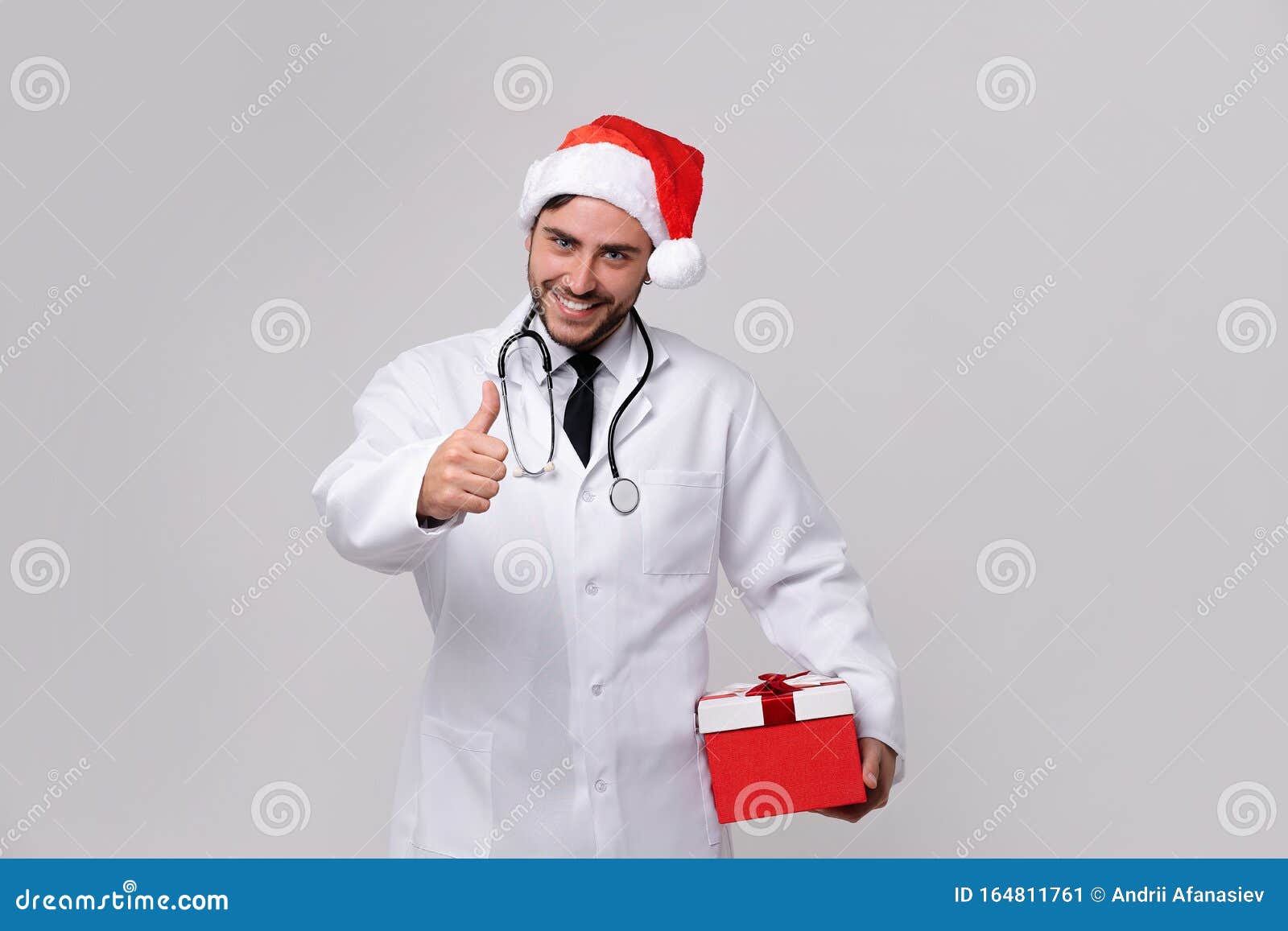 young handsome doctor in white uniforme and santa claus hat standing in studio on red background smile and finger in camera