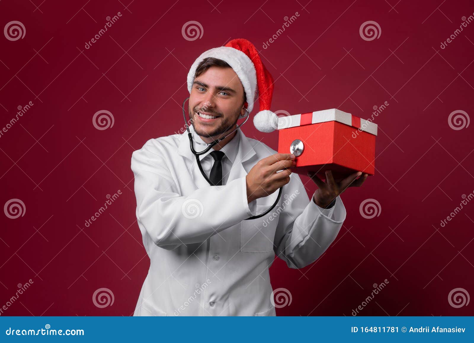 young handsome doctor in white uniforme and santa claus hat standing in studio on red background smile and finger in camera