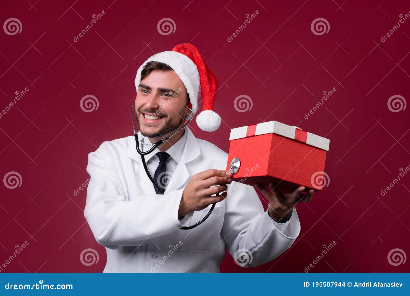 young handsome doctor in white uniforme and santa claus hat standing in studio on red background smile and finger in camera